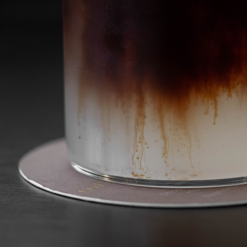 A close-up of a frosted glass tumbler with a dark brown gradient wash near the top, resting on a round metallic coaster against a dark background.