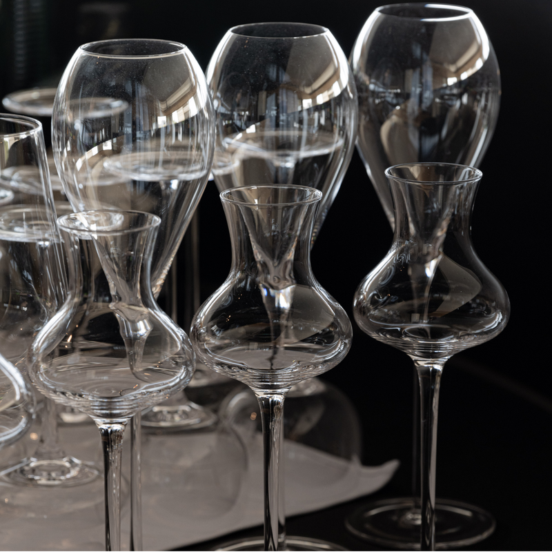 A group of clear glass wine glasses with different shapes and lengths, arranged closely together on a dark reflective surface.