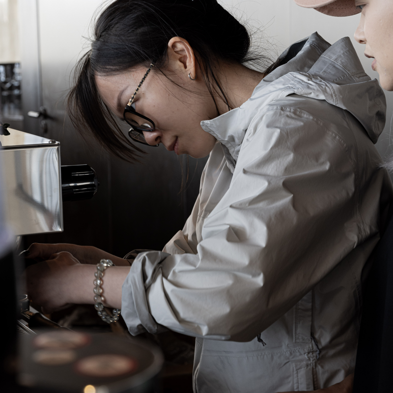 A woman wearing a light gray hooded jacket and glasses leans over a counter in a cafe setting, with another person partially visible on the right.
