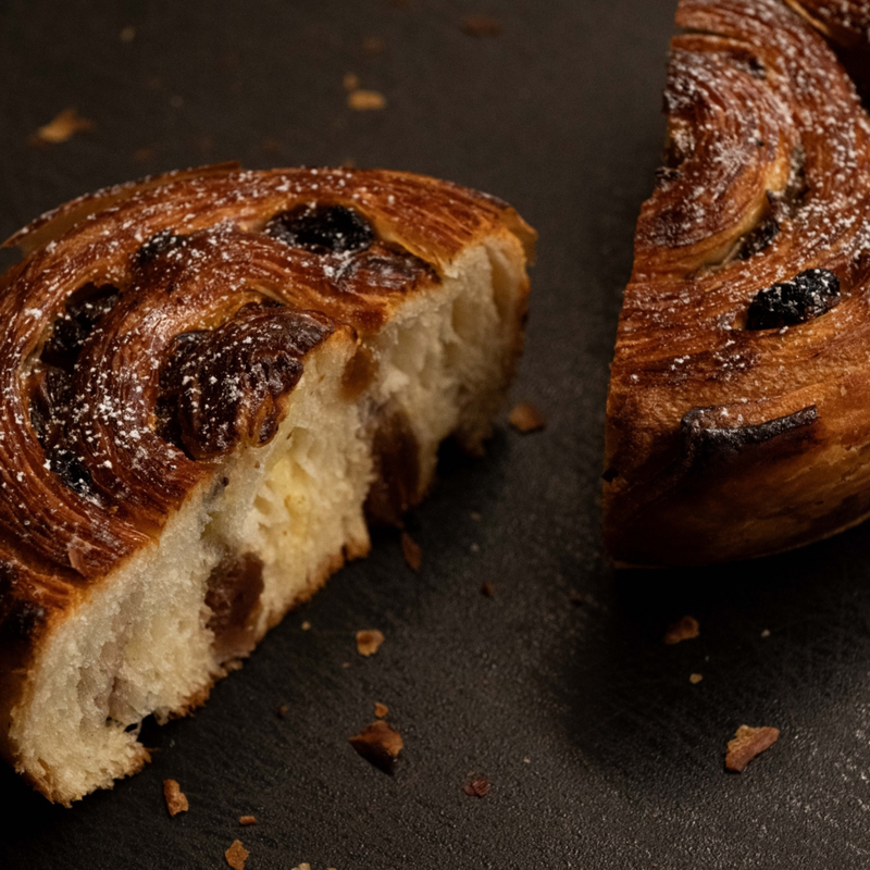 A close-up of a baked pastry or bread roll cut into pieces, showing a glossy golden-brown flaky crust with dark chocolate swirls and a soft cream-colored interior, set on a dark surface.