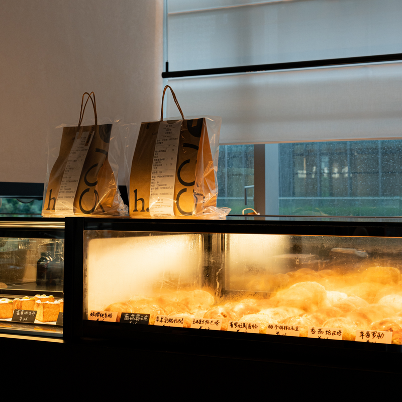 Two branded paper takeaway bags with brown handles sit on top of a bakery display case in a cafe or restaurant. The case is illuminated from within, showing pastries and bread, with a window and dark interior visible in the background.