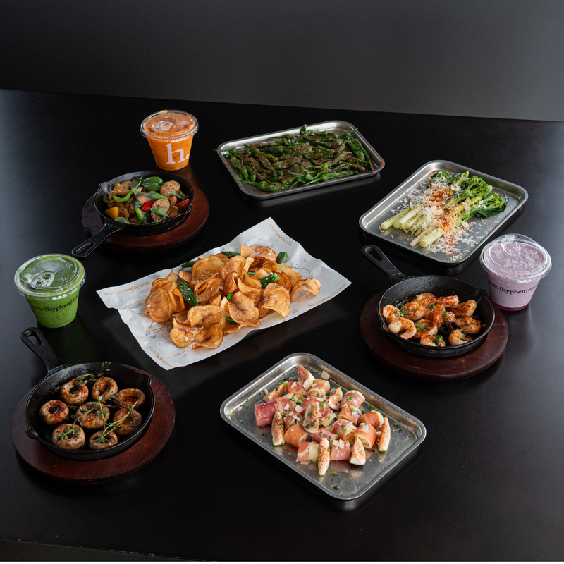 A variety of prepared foods and drinks arranged on a black table, including several cast-iron skillet dishes, two metal trays, a paper-lined platter of chips, and two plastic cups with lids.