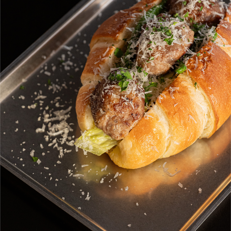 A close-up of a gourmet stuffed pastry or bread roll on a dark baking tray, filled with meatballs topped with grated cheese and chopped green herbs, with a piece of lettuce visible.
