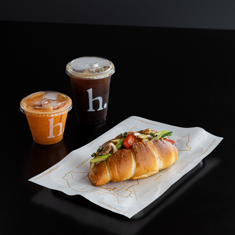 A croissant sandwich with avocado, tomato, and mushroom filling on a branded paper plate, alongside two iced coffee drinks with a clear plastic lid and a cup with a white 'h.' logo, on a black background.