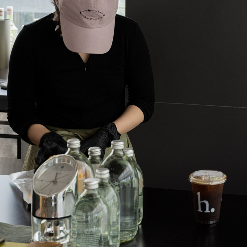 A person in a black long-sleeve top, black gloves, and a light pink cap is standing behind a counter with several clear glass bottles and a coffee cup with a visible 'h.' logo.