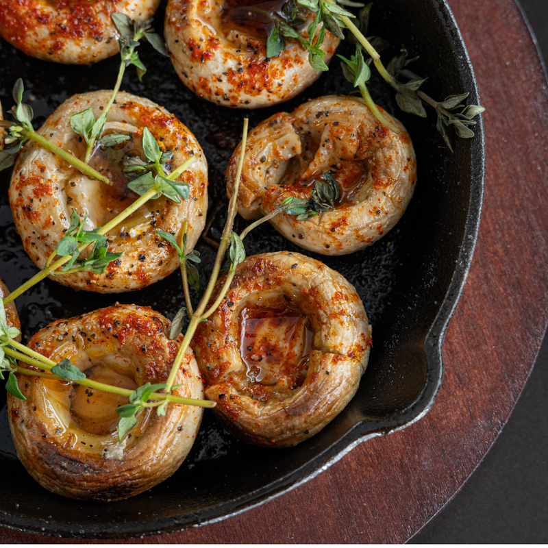 Six baked mushroom caps arranged in a black cast-iron skillet, topped with melted butter, herbs, and red pepper seasoning, photographed from above on a dark wooden surface.