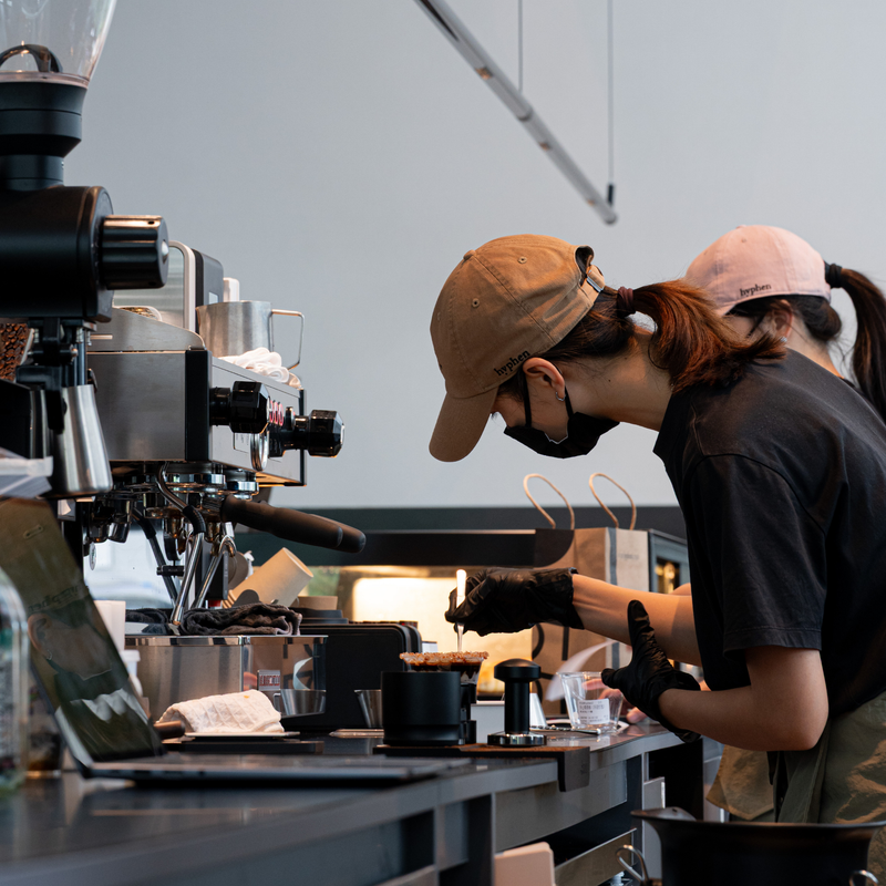 A barista in a tan cap, black face mask, and black shirt prepares a drink behind a coffee station, with another person partially visible behind them in a cafe setting.