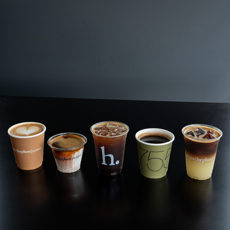 Five takeaway coffee cups from Hyphen Juice arranged in a row on a dark table against a dark gray wall. The cups include different coffee preparations: a latte, a small espresso shot, an iced coffee, a black coffee cup with a '75' design, and an iced coffee with a yellow gradient.