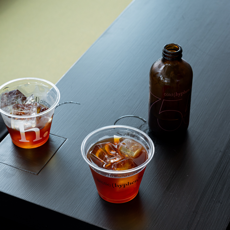 Two clear plastic cups containing an amber drink with ice, a dark brown bottle, and a small coaster on a dark tabletop.