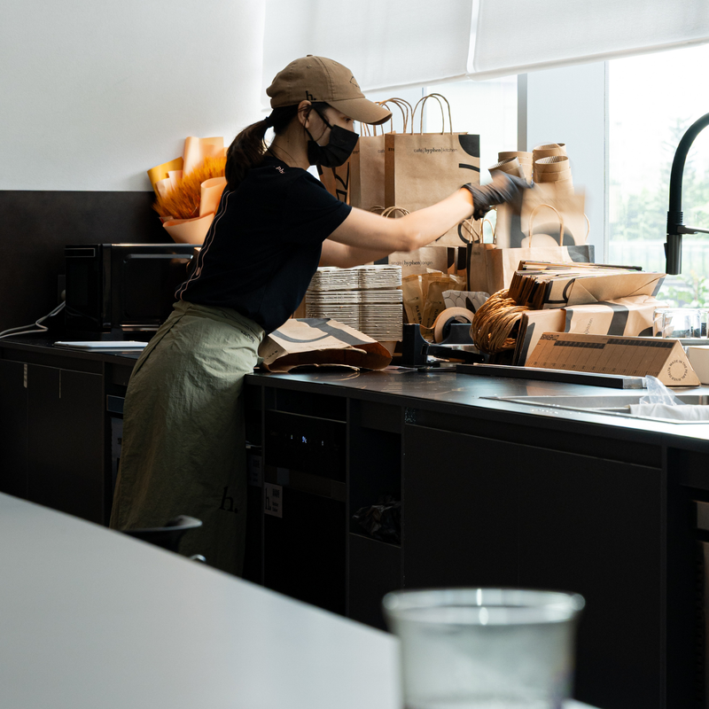 A person in a black shirt, khaki pants, and a tan cap packs paper bags at a counter in a restaurant or cafe kitchen area.