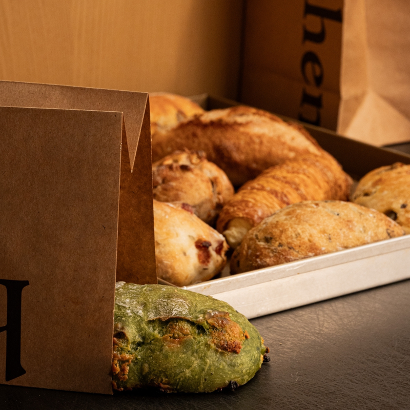 A tray of assorted baked pastries and breads, including croissants, scones, and a green savory bread, with brown paper packaging in the foreground and background.