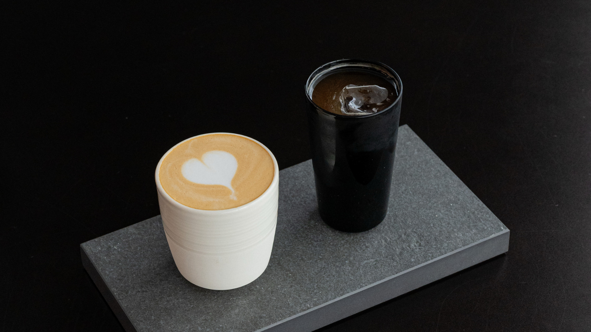 A ceramic coffee cup with a heart-shaped latte art design sits next to a black glass tumbler with ice on a dark gray rectangular tray against a black background.