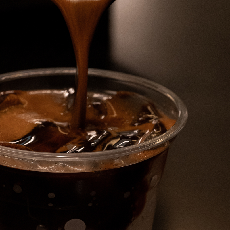 A close-up shot of a thick, glossy brown sauce or syrup being poured from above into a clear plastic cup filled with iced coffee and ice.