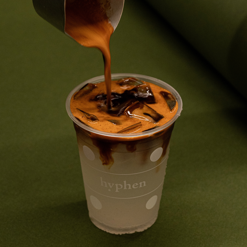 A clear plastic cup with white polka dots and the word 'hyphen' printed on the front, filled with an iced coffee drink and topped with a pour of espresso, set against a dark olive green background.