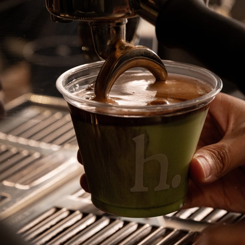 A hand holds a clear plastic cup with a green base and a large gray letter 'h.' while espresso pours into the cup from an espresso machine.