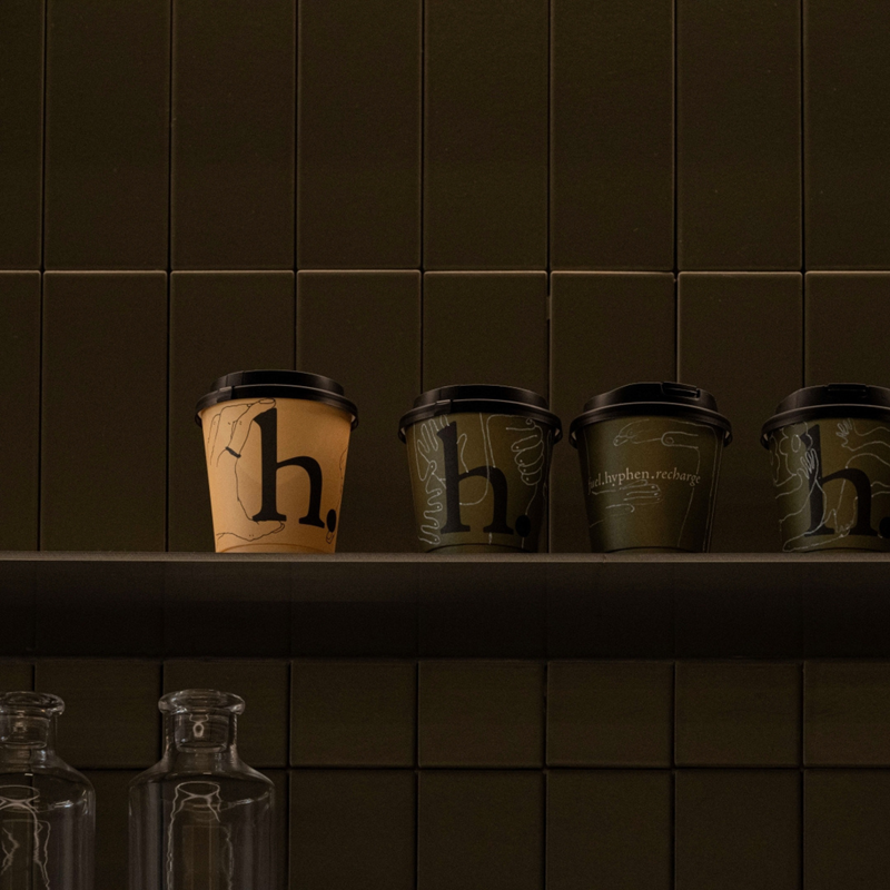 Four takeaway coffee cups with lids lined up on a dark shelf against a dark tiled wall. Three cups are olive green with white line art and the letter 'h.