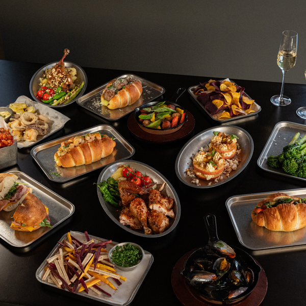 An overhead tabletop display of multiple prepared food dishes arranged on metal trays and plates, including wraps, pasta, grilled chicken, shrimp, broccoli, mussels, and a glass of white wine on a dark table.