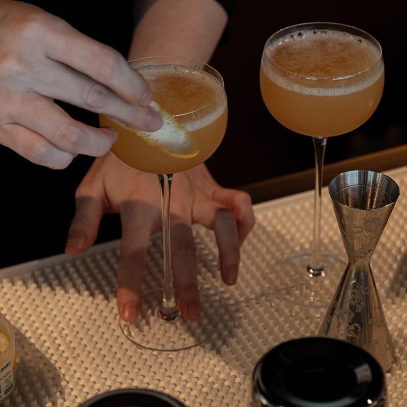 A close-up of a bartender's hands preparing a cocktail in a dimly lit bar setting. One hand holds a stemmed glass filled with an amber-orange foamy drink and garnishes it with a lemon twist, while another identical glass stands nearby.