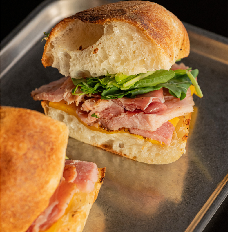 A close-up of a gourmet sandwich on a metal tray, featuring a rustic ciabatta bread, melted cheese, sliced ham, and fresh green arugula.