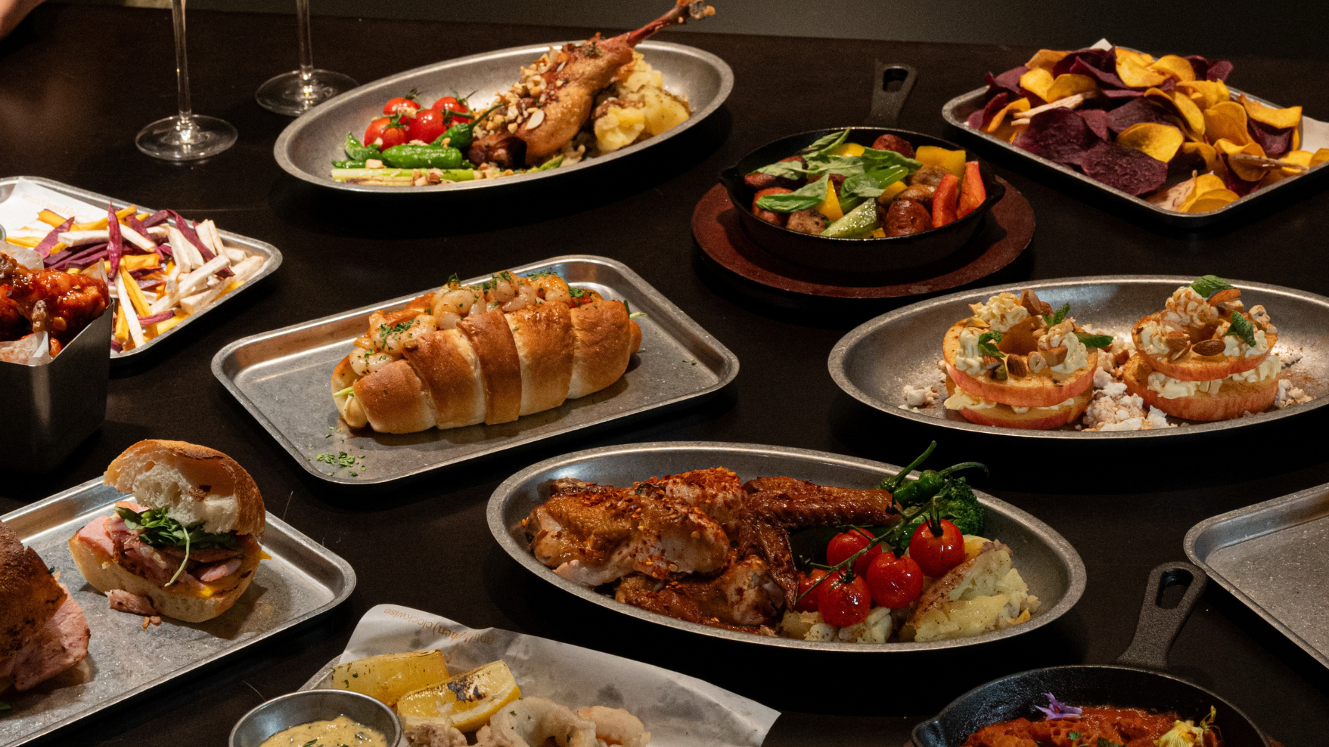 A table laden with assorted prepared foods and snacks, including grilled meats, bread rolls, chips, and salads, photographed in a warm indoor setting.