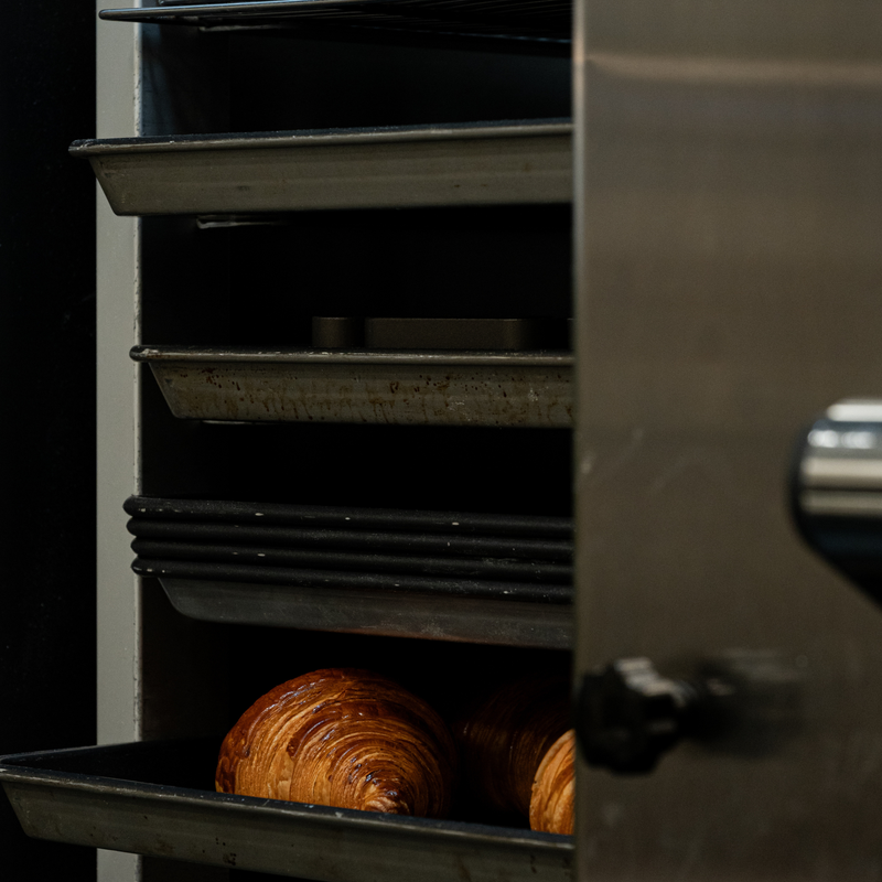A stainless steel commercial bread oven with trays inside, including two golden-brown croissants on the bottom shelf.