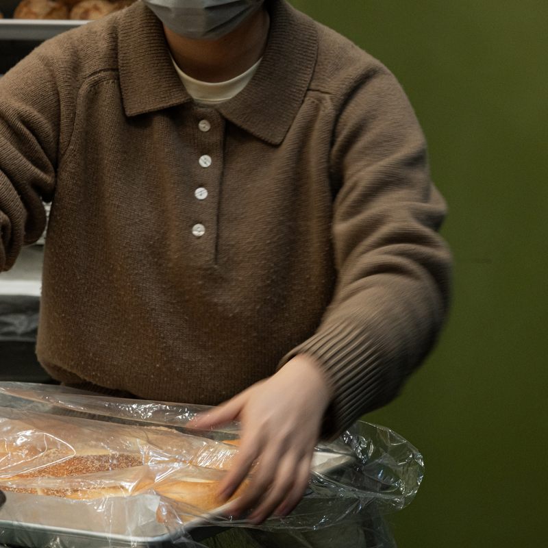 A person wearing a brown knit polo sweater and a gray face mask is shown from the chest down, reaching toward packaged food on a tray in what appears to be a kitchen or food prep area.