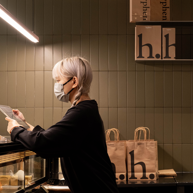 A person with short silver hair, wearing a black long-sleeve top and a light gray face mask, stands at a counter in a cafe or restaurant, holding a tablet. The background features a dark olive-green tiled wall with shelves holding books and branded paper bags with the logo 'h.