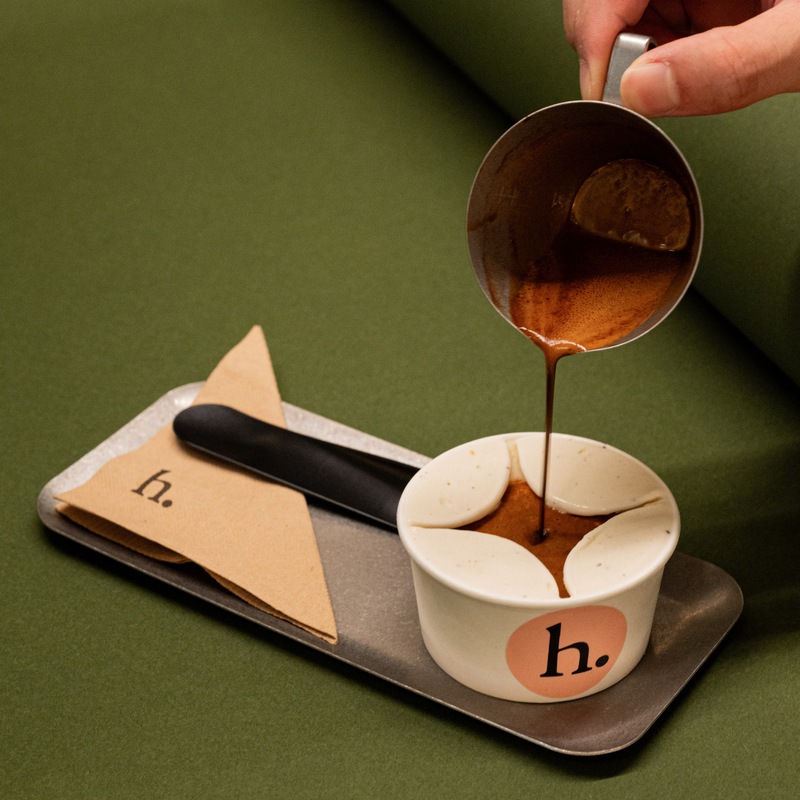 A hand pours espresso into a white paper cup with a pink circular logo, set on a metal tray with a black spatula and a brown paper note marked 'h.' against an olive green background.
