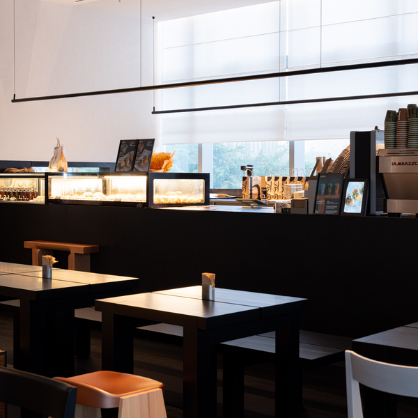 A modern cafe interior with dark tables, a black counter, and a display case filled with pastries. A coffee machine is visible on the right, and large windows with white blinds are in the background.