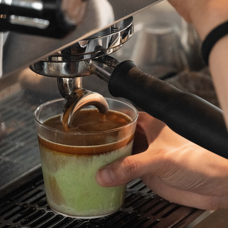 A close-up of a hand holding a clear glass cup under a metal espresso portafilter with a black handle, pouring espresso into a layered drink that shows a pale green liquid beneath a brown top layer.