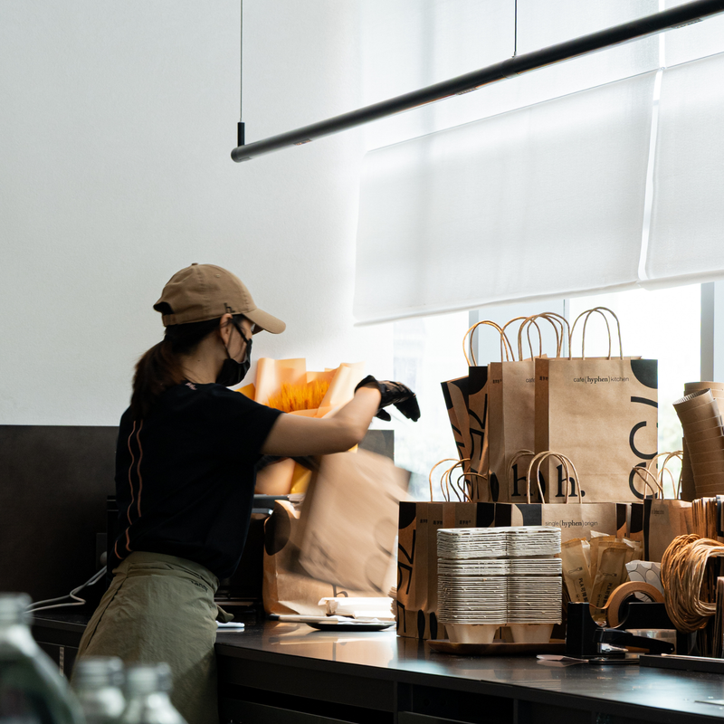 A hyphen. team member packing an order at the counter, beside stacked kraft takeaway bags printed cafe (hyphen) kitchen and single (hyphen) origin