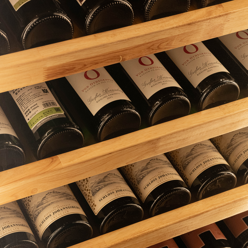 A close-up of a wooden wine rack holding several dark glass wine bottles with cream labels. The wood shelves are arranged diagonally, and the labels show wine names such as Schloss Johannisberg.